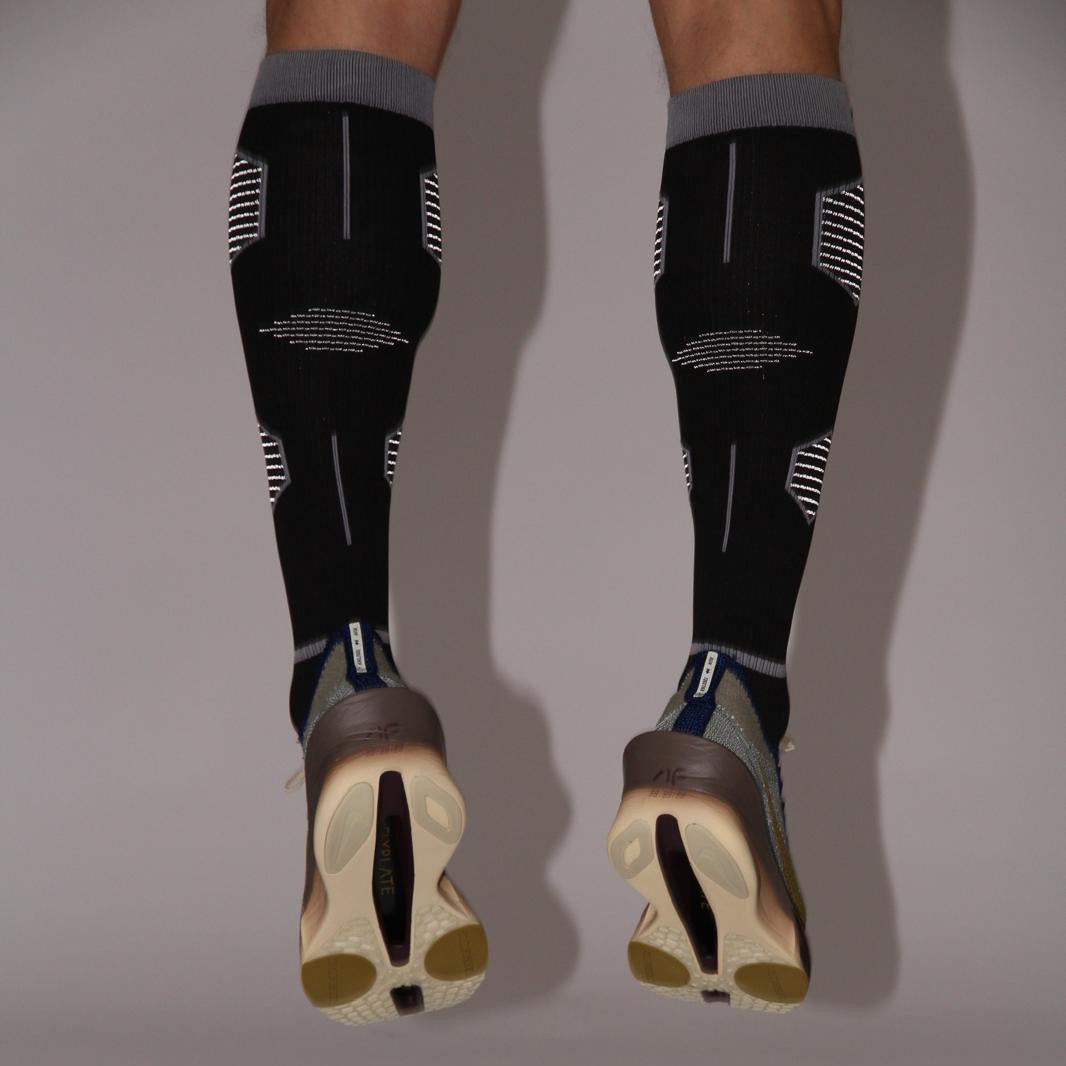 Black compression socks with gray tops worn by a person on a plain background
